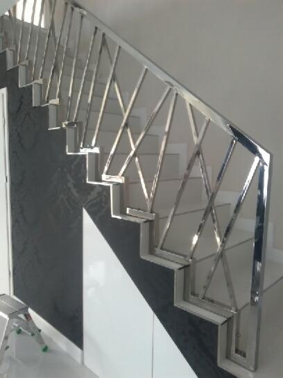 Nierdzewna balustrada schodowa o geometrycznym wzorze, widoczna z dołu, z fragmentem drabiny malarskiej w lewym dolnym rogu.