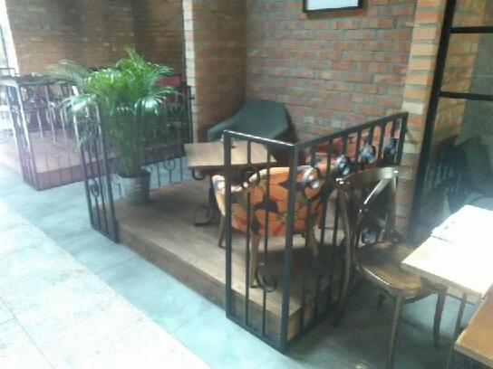 Metalowa balustrada z dekoracyjnymi elementami otaczająca podest z rośliną doniczkową w kawiarni o ceglanych ścianach.