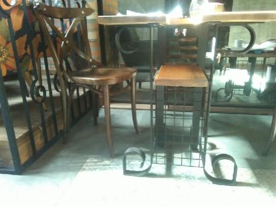 Drewniane krzesło z krzyżowym oparciem i metalowa konstrukcja stołka barowego z siedziskiem z jasnego drewna, oba elementy w surowym wnętrzu z fragmentem balustrady z metalowymi zdobieniami.