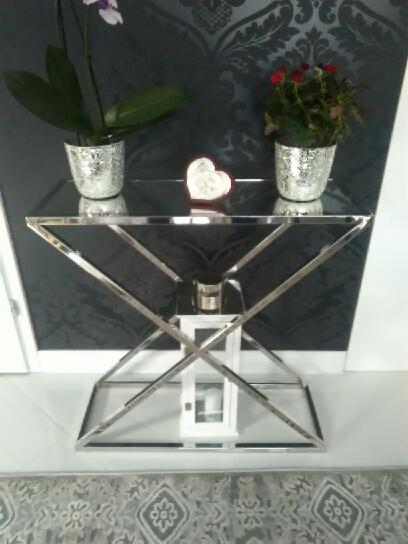 Szklany stolik konsola z chromowanymi nogami w kształcie litery X, na blacie dwie doniczki z kwiatami i dekoracja w kształcie serca, pod blatem biała latarenka.