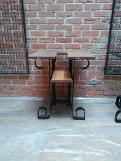 Stolik barowy z drewnianym blatem i metalową konstrukcją w stylu industrialnym, na tle ściany z czerwonej cegły i metalowych krat.