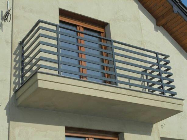 Nowoczesna, szara balustrada balkonowa z poziomych profili stalowych, zamontowana na betonowej płycie balkonu, widok z dołu na tle elewacji budynku.