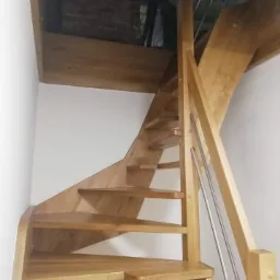 Spiralne, jasne, drewniane schody z metalową balustradą wewnątrz pomieszczenia, widoczne z dołu, prowadzące na antresolę z ceglanym wykończeniem.