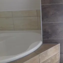 Narożna kabina prysznicowa z jasnymi płytkami na ścianach i ciemnymi, drewnopodobnymi płytkami na ścianie obok okna, wykończona listwą przy podłodze z płytek.