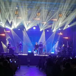 Zespół muzyczny na scenie podczas koncertu, oświetlony reflektorami, widownia nagrywa występ telefonami.