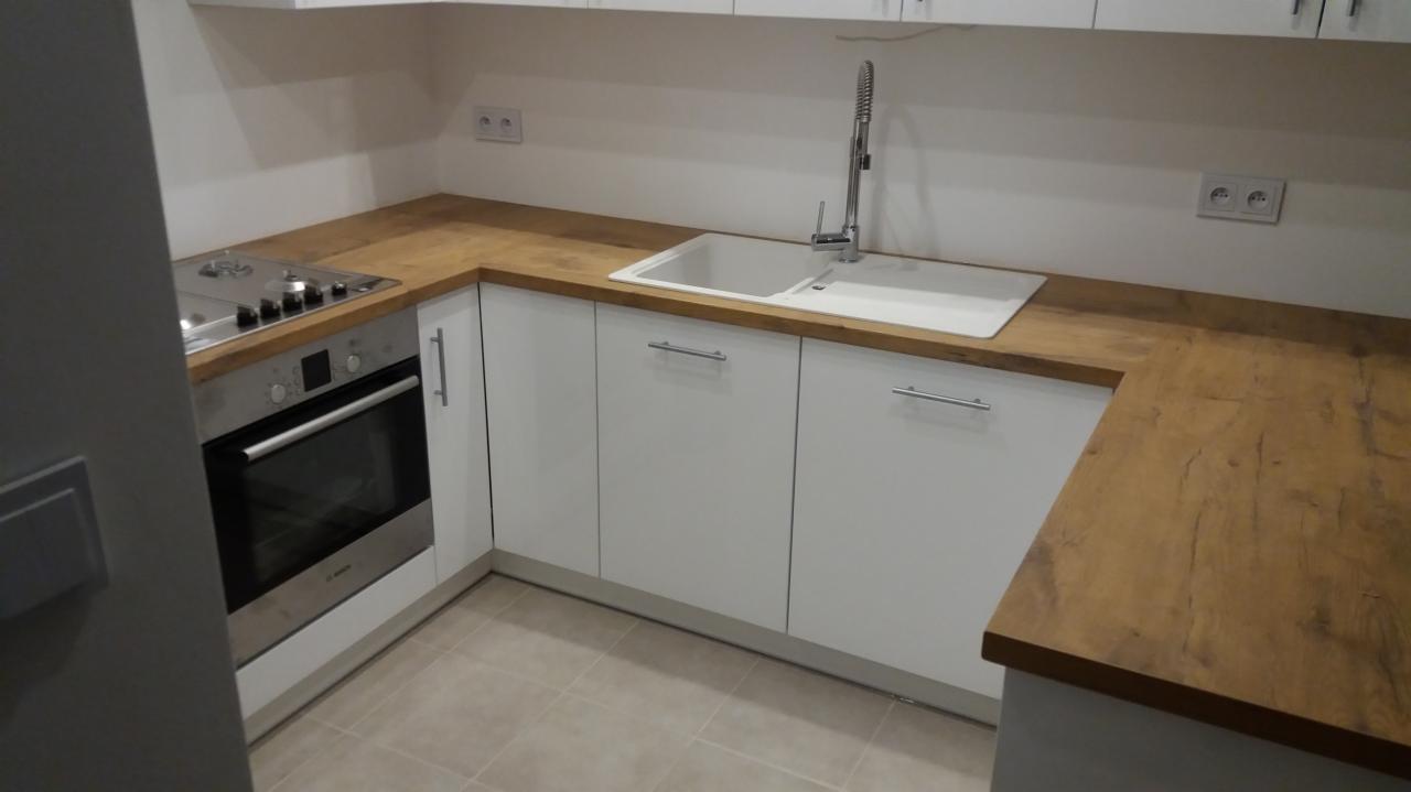 Jasna kuchnia z białymi szafkami i blatem imitującym drewno, widoczny zlew, bateria i płyta grzewcza z piekarnikiem.