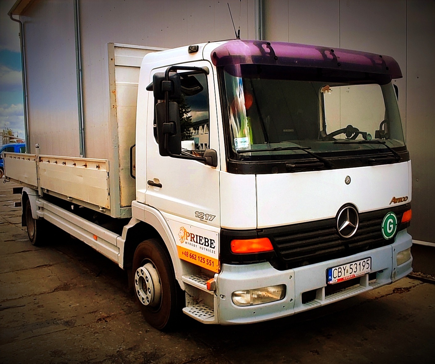 Biały samochód ciężarowy Mercedes Atego 1217 z zabudową skrzyniową i pomarańczową naklejką reklamową firmy Priebe Wyroby Hutnicze na błotniku, zaparkowany na betonowym placu.