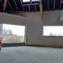 Wnętrze w trakcie prac tynkarskich, z widoczną maszyną do tynkowania w kolorze pomarańczowym, świeżo otynkowane ściany w odcieniach szarości, surowy strop z oknami dachowymi i widokiem na zewnątrz...