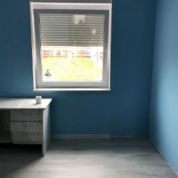 Pomalowany na błękitno pokój z jasną podłogą i oknem z roletami, biurko z szufladami przy ścianie, widok na zewnątrz przez okno.