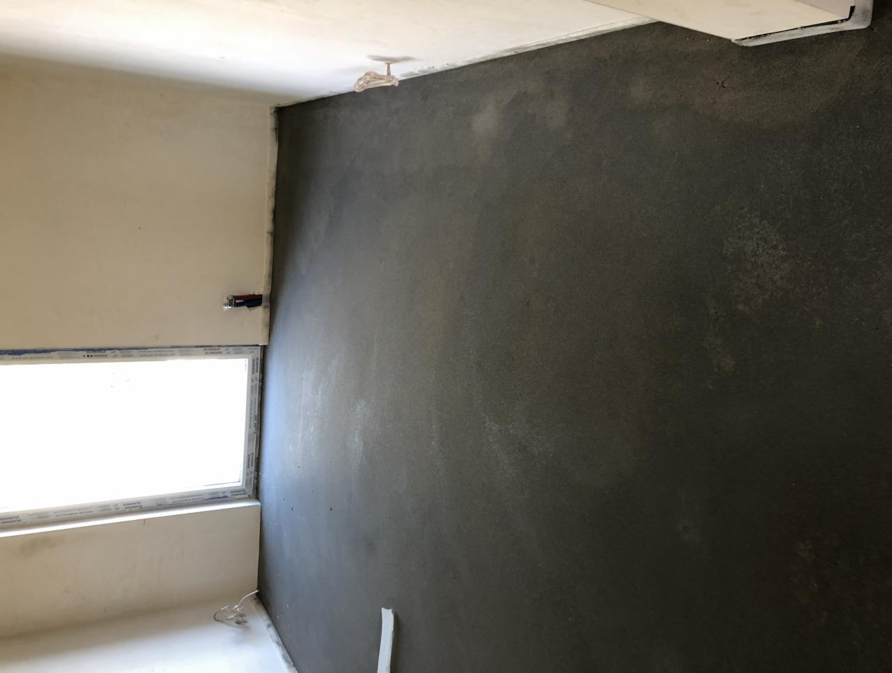 Świeżo nałożona, ciemnoszara warstwa tynku na ścianie w pomieszczeniu z oknem. Widoczny fragment sufitu z wystającym elementem instalacji.
