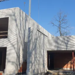 Wznoszenie dwukondygnacyjnego budynku z białej cegły, widoczny pracownik w kasku oraz częściowo wykończone otwory okienne i drzwiowe z czerwonej cegły, na zewnątrz drzewa i niebieskie niebo.