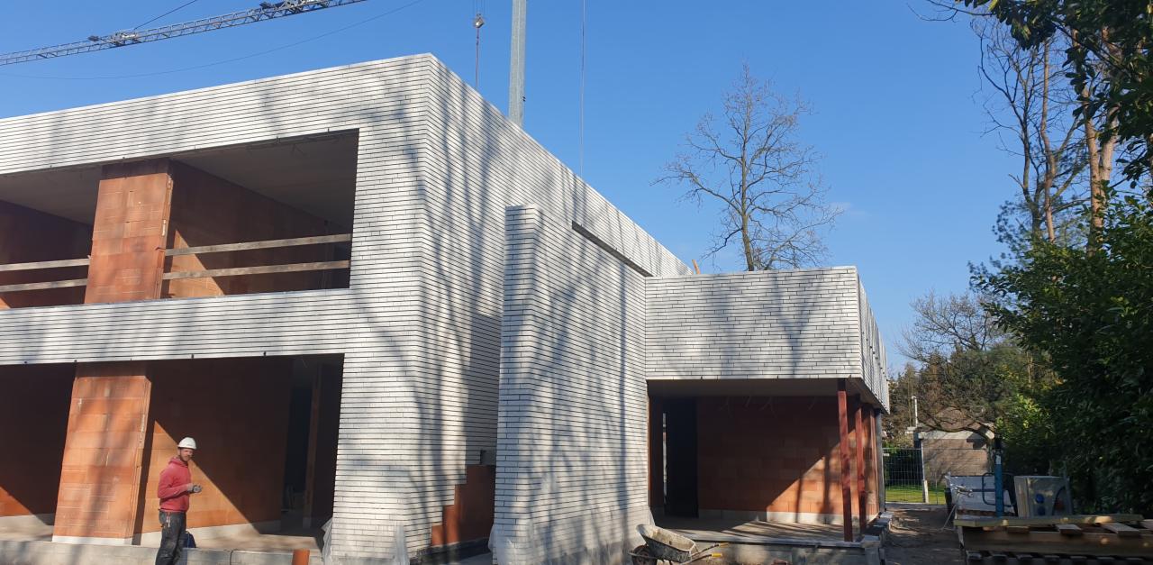 Wznoszenie dwukondygnacyjnego budynku z białej cegły, widoczny pracownik w kasku oraz częściowo wykończone otwory okienne i drzwiowe z czerwonej cegły, na zewnątrz drzewa i niebieskie niebo.