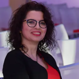 Portret uśmiechniętej kobiety w okularach, ubrana w czerwoną bluzkę i czarny żakiet, siedzącej na tle białych krzeseł w sali konferencyjnej.