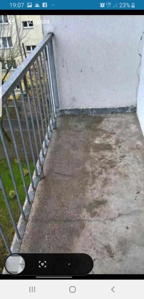 Zaniedbany balkon z betonową posadzką, widoczne plamy i zabrudzenia, metalowa balustrada z pionowymi szczeblami, ściana z widocznymi śladami wilgoci i pęknięciem, widok na zieleń poza balkonem.
