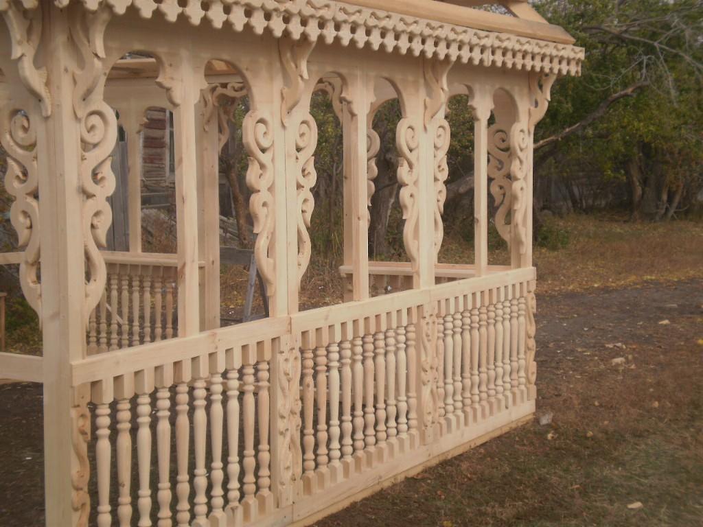 Surowa drewniana altana z ozdobnymi wycięciami i balustradą, w trakcie budowy na tle jesiennego krajobrazu.