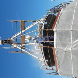 COATING POLAND - Prace konserwacyjne na statku: robotnik w kombinezonie, na rusztowaniu, aplikuje strumień czyszczący na kadłub statku, widoczne elementy antenowe i osłona z folii, błękitne niebo w tle.