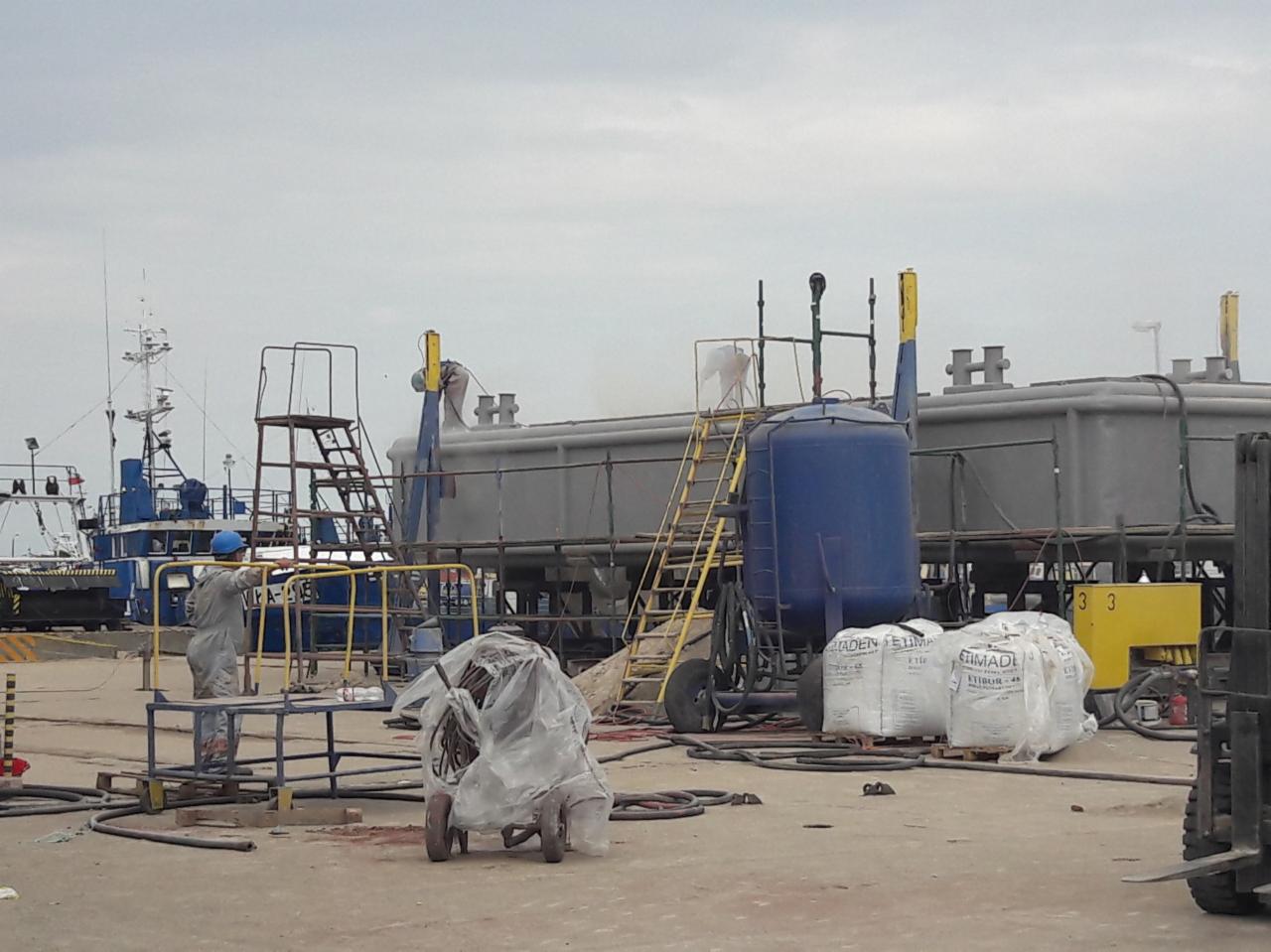 Prace przygotowawcze do malowania hydrodynamicznego dużego zbiornika na terenie portu, widoczni pracownicy w kombinezonach ochronnych i hełmach, na drugim planie statki.