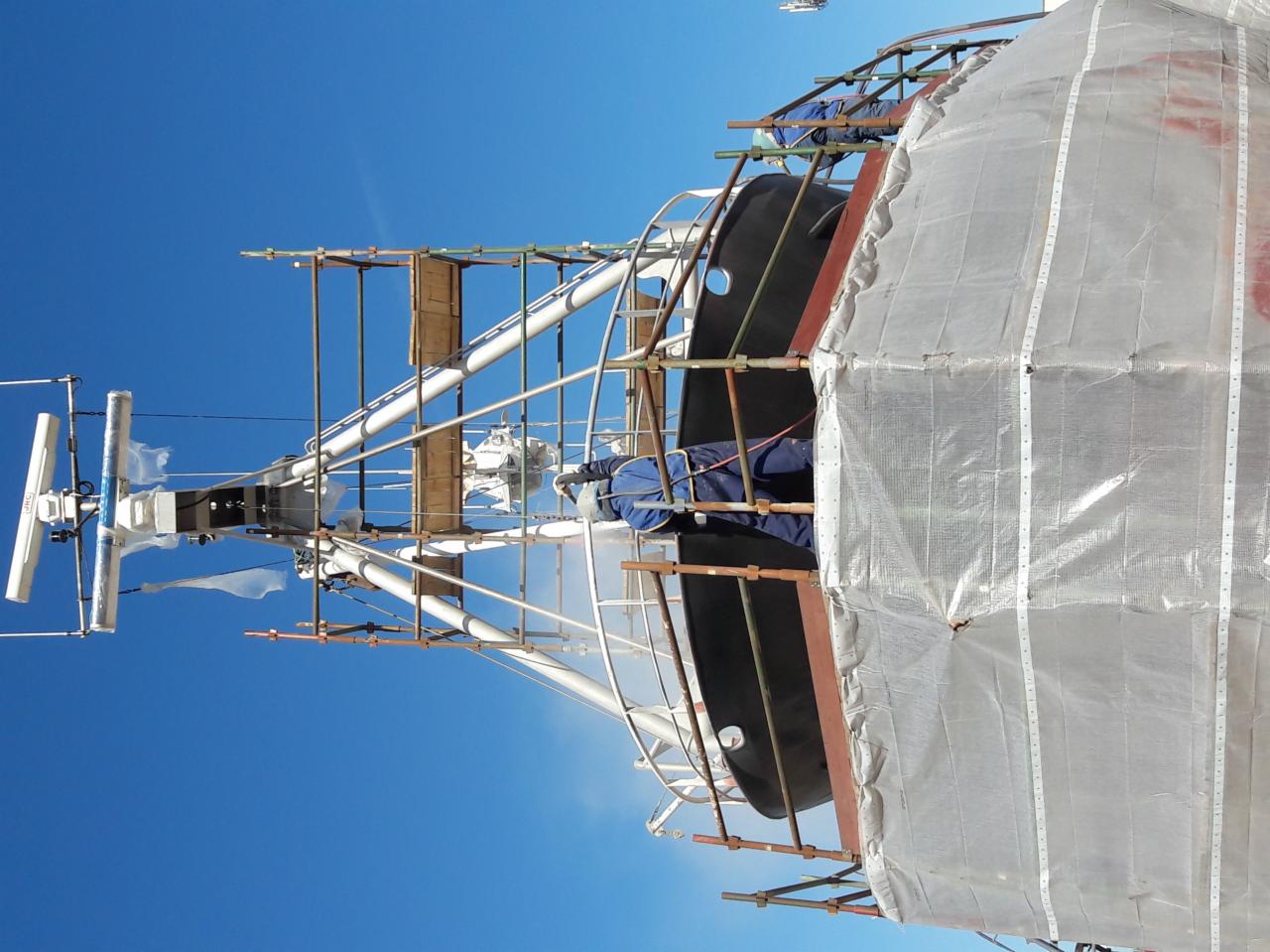 Prace konserwacyjne na statku: robotnik w kombinezonie, na rusztowaniu, aplikuje strumień czyszczący na kadłub statku, widoczne elementy antenowe i osłona z folii, błękitne niebo w tle.