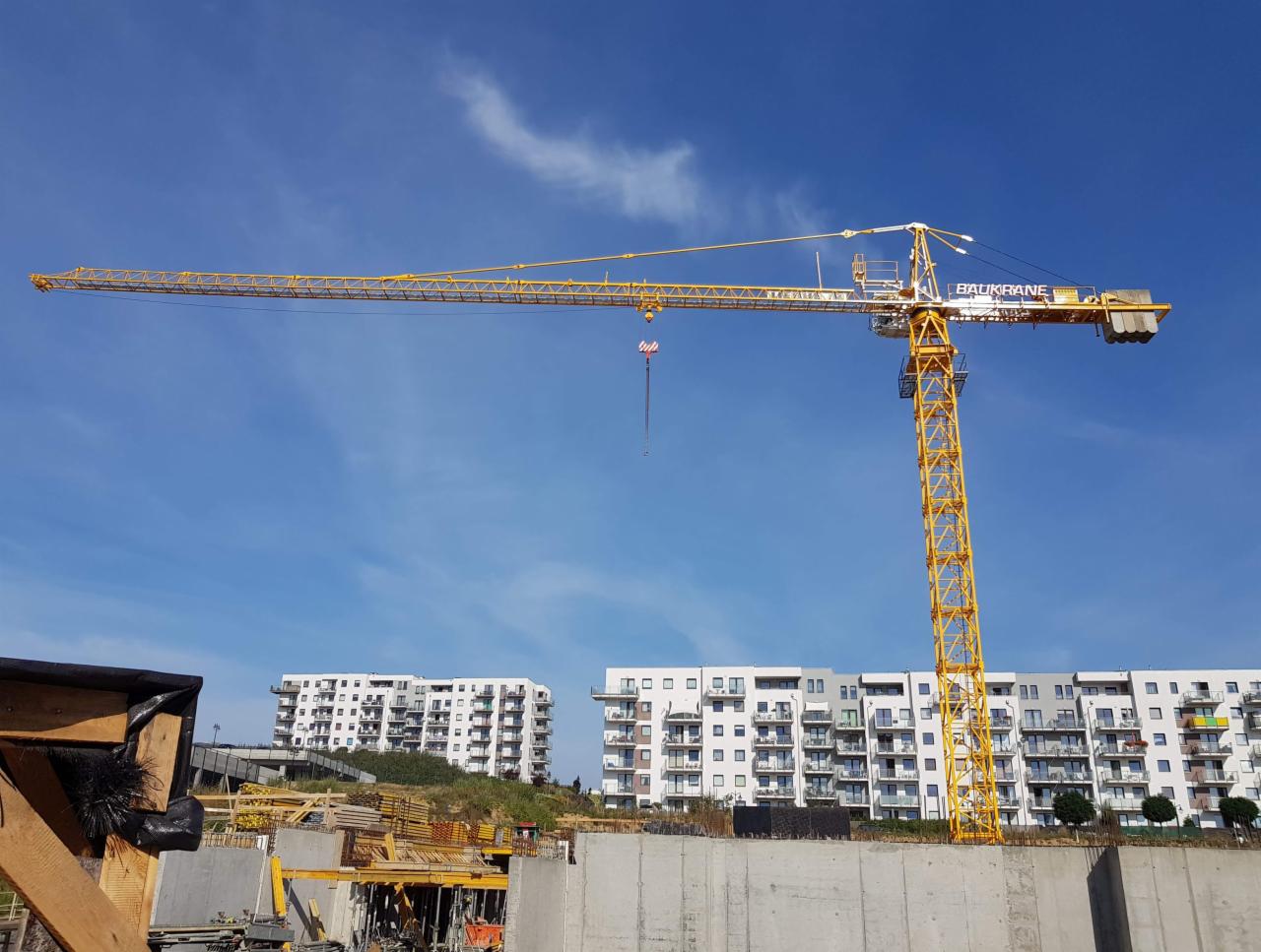 Żółty dźwig budowlany na tle błękitnego nieba i budynków mieszkalnych w Gdyni, widoczny plac budowy z elementami szalunków.