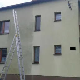 Dwukondygnacyjny dom z częściowo odnowioną elewacją, widoczne różnice w kolorze i czystości, oparta o ścianę drabina aluminiowa, w tle plac zabaw dla dzieci.