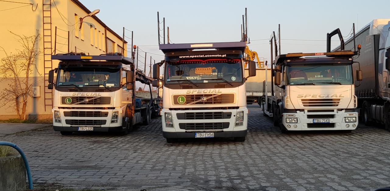 Trzy białe ciężarówki marki Volvo Special zaparkowane na brukowanym placu, przygotowane do załadunku lub rozładunku, z widocznymi platformami do przewozu samochodów.