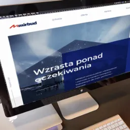 Ekran komputera iMac wyświetlający stronę internetową firmy Mirbud z hasłem 'Wzrasta ponad oczekiwania', obok klawiatura i dłoń obsługująca mysz.