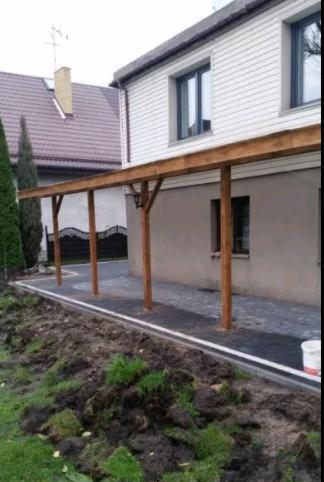 Wykończona ścieżka z ciemnej kostki brukowej przy domu z drewnianą konstrukcją zadaszenia, widoczne niedokończone prace ziemne przy trawniku.