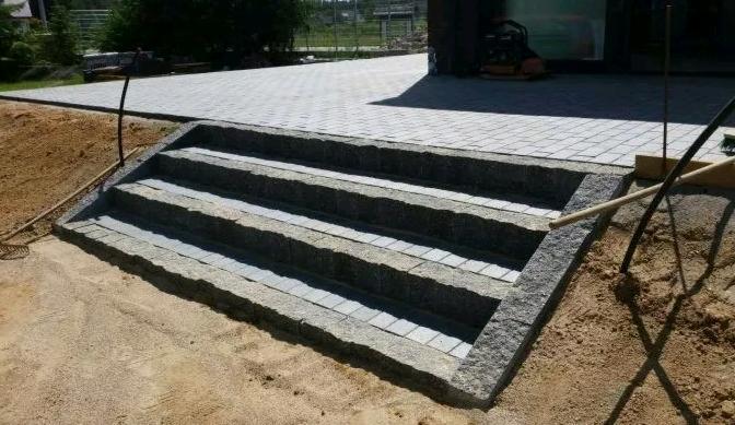 Granitowe schody wejściowe z kostki, prowadzące do domu z brukowaną ścieżką, w trakcie prac wykończeniowych.