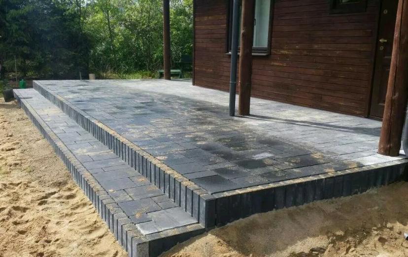 Nowo ułożony taras z ciemnoszarej kostki brukowej, z dwustopniowymi schodami i obrzeżami, przylegający do drewnianego budynku z widocznymi słupami werandy i piaskiem wokół.