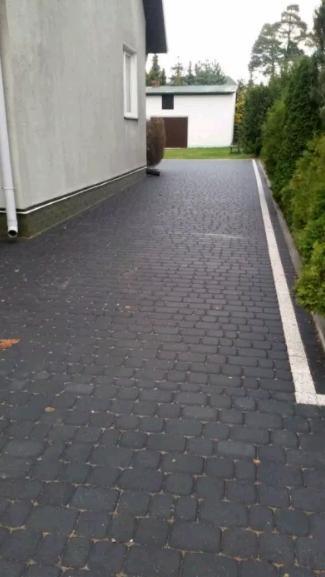 Ciemnoszara kostka brukowa ułożona na podjeździe w Augustowie, z widocznym obrysem białej farby na krawędzi, przylegająca do budynku z szarą elewacją i rynną.