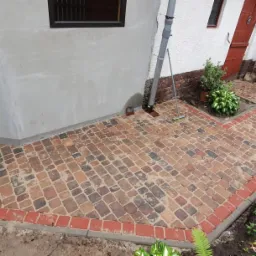 Ścieżka z kostki brukowej w odcieniach brązu i czerwieni, obrzeże z czerwonej kostki, prowadząca wzdłuż budynku z białymi ścianami i brązowymi oknami, widoczne rośliny i trawnik.