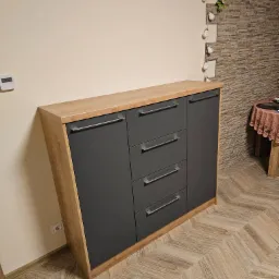 Nowoczesna komoda z szufladami i szafką, wykończona drewnem i szarym matem, na tle beżowej ściany i podłogi w jodełkę. Widoczny fragment dekoracji ściennej.