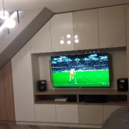 Biała zabudowa meblowa pod schodami z szafkami, półkami i miejscem na TV. Nowoczesne oświetlenie schodów i sufitu.