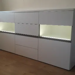 Biała komoda z szufladami i szafkami, podświetlana od wewnątrz, stoi na drewnianej podłodze w jasnym pomieszczeniu. Minimalistyczny design.