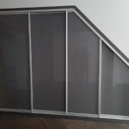 Szafa wnękowa z przesuwnymi drzwiami, dopasowana do skosu sufitu. Szare fronty, srebrne ramy, widoczna ceglana ściana w tle. W lustrze odbija się osoba robiąca zdjęcie.