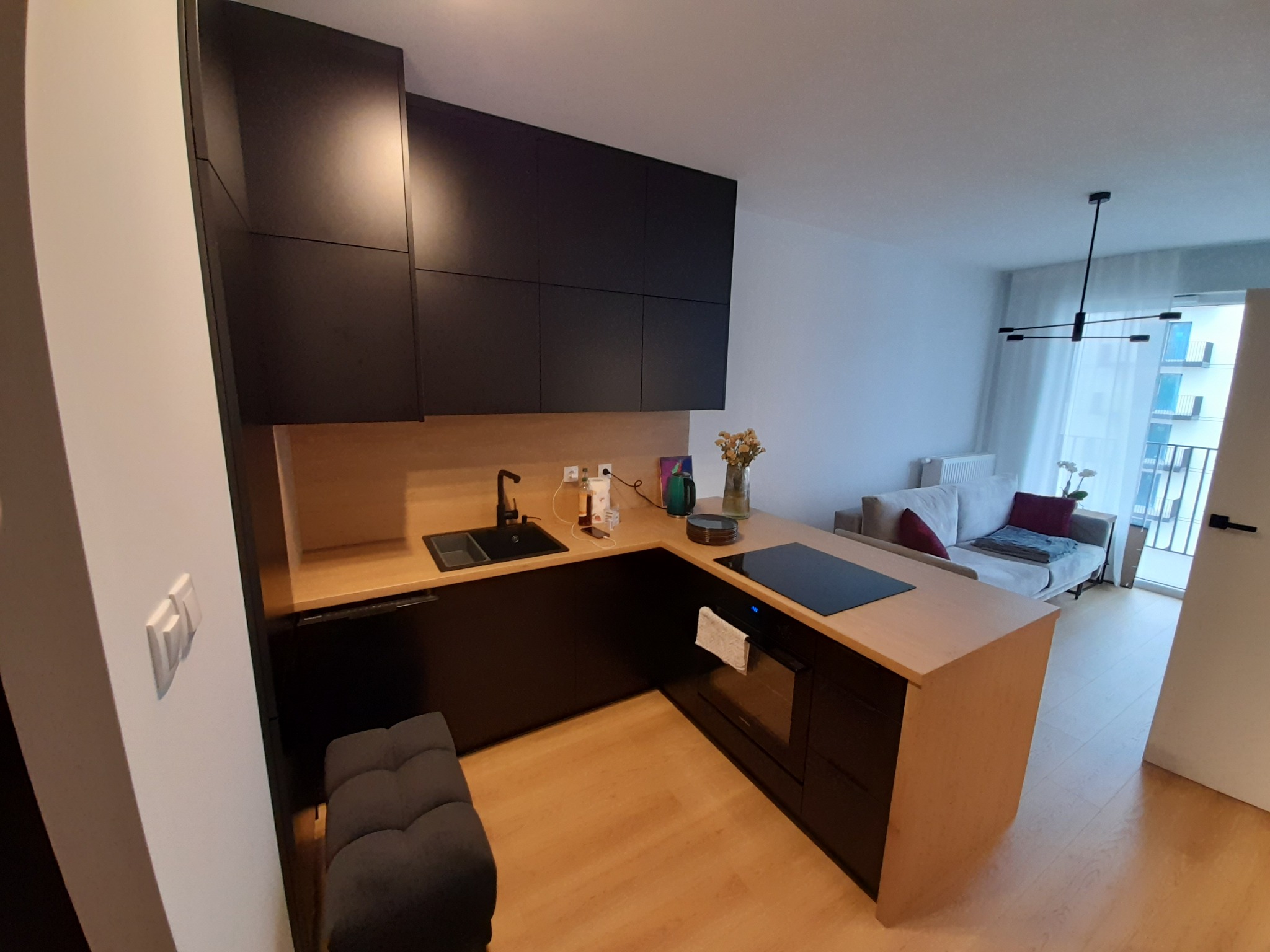 Nowoczesna kuchnia z czarnymi szafkami i drewnianym blatem, minimalistyczny design, widok na salon z kanapą i balkonem, jasne wnętrze.