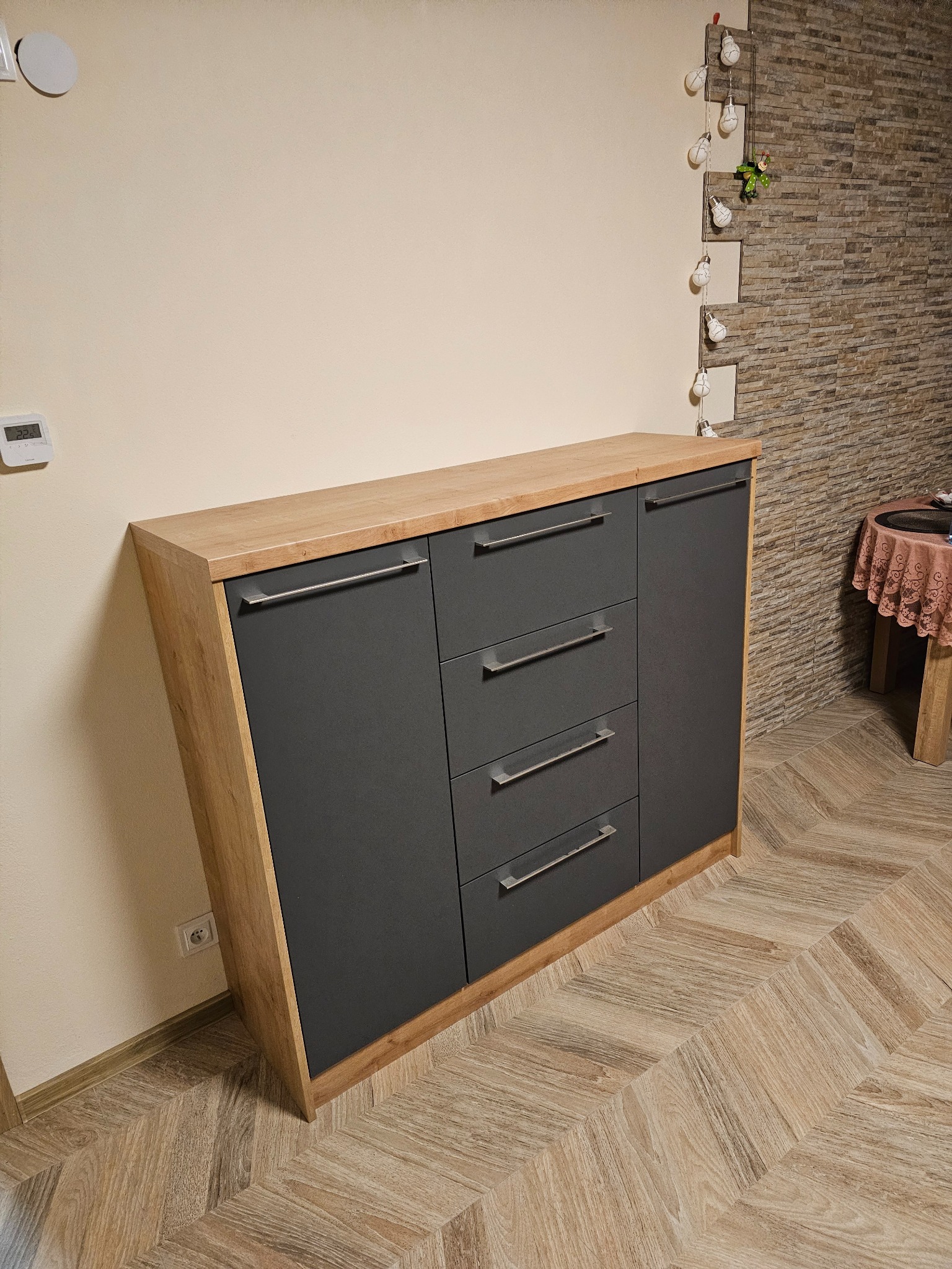 Nowoczesna komoda z szufladami i szafką, wykończona drewnem i szarym matem, na tle beżowej ściany i podłogi w jodełkę. Widoczny fragment dekoracji ściennej.
