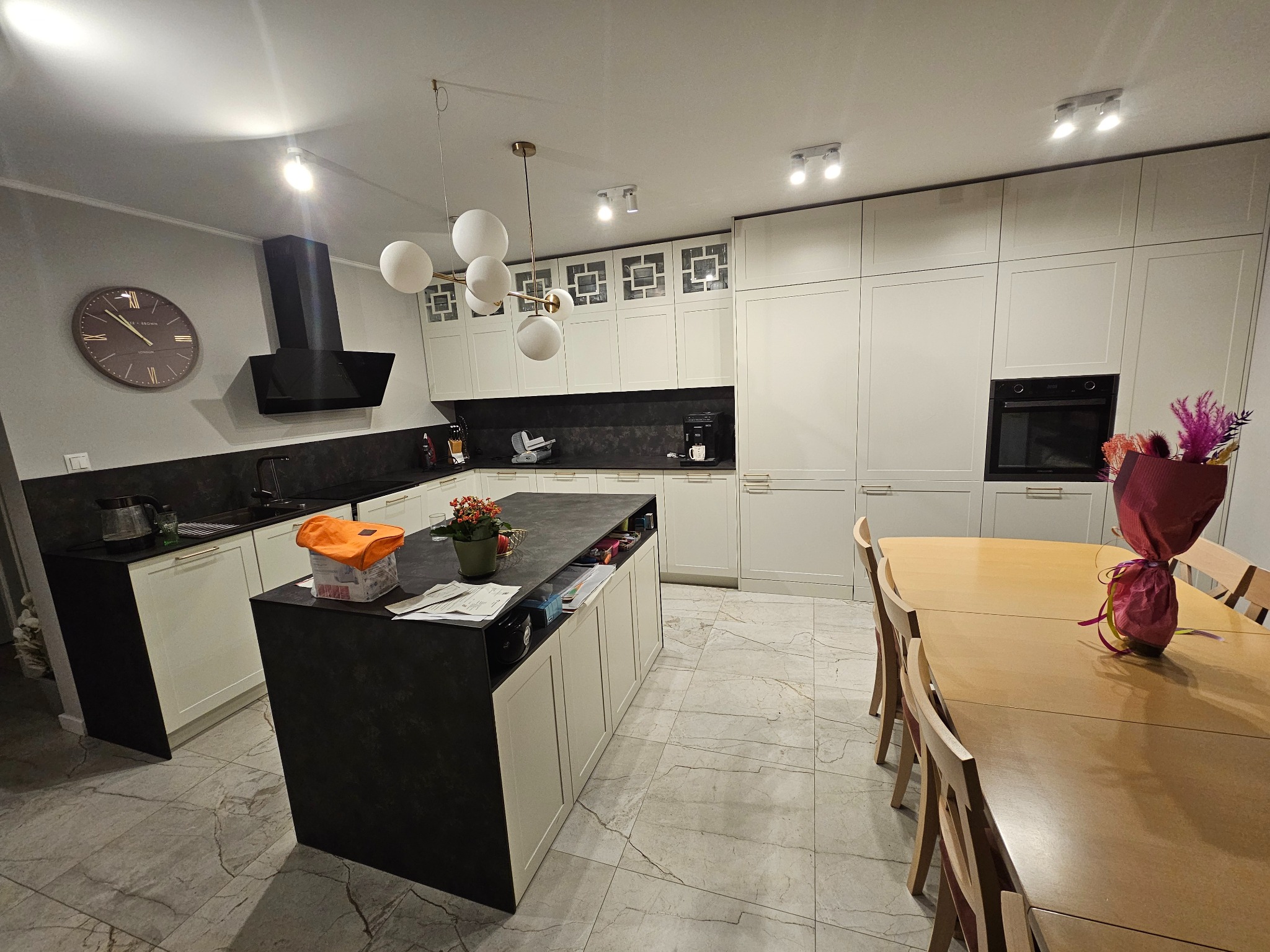 Jasna kuchnia z wyspą i stołem jadalnym. Białe szafki, czarny blat wyspy i okap. Na wyspie kwiaty i akcesoria. Marmurowa podłoga. Nowoczesne oświetlenie.
