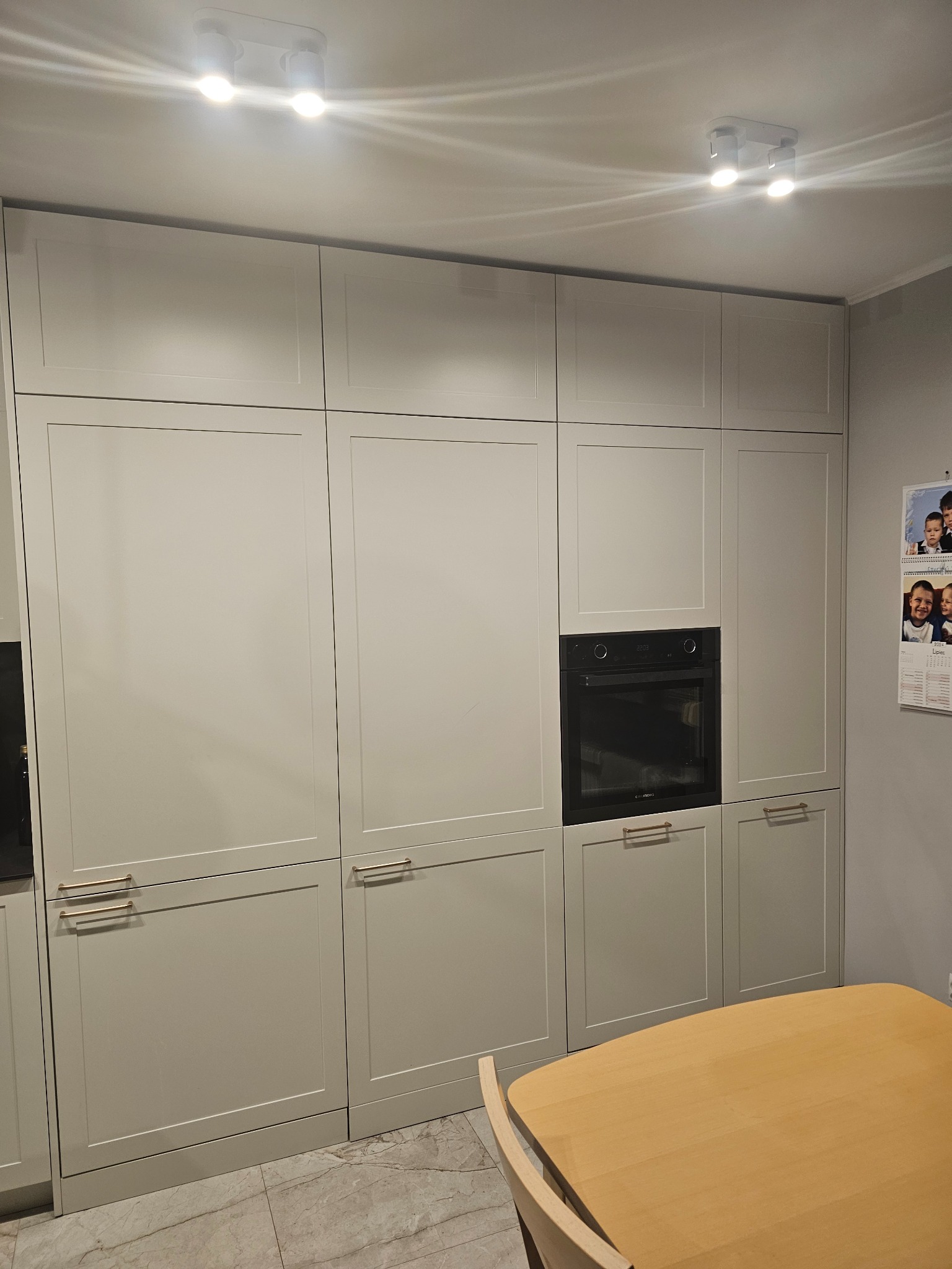 Jasnoszare szafki kuchenne na wymiar z piekarnikiem w zabudowie, widoczne w Piotrków Trybunalski. Proste uchwyty, marmurowe płytki na podłodze i drewniany stół.