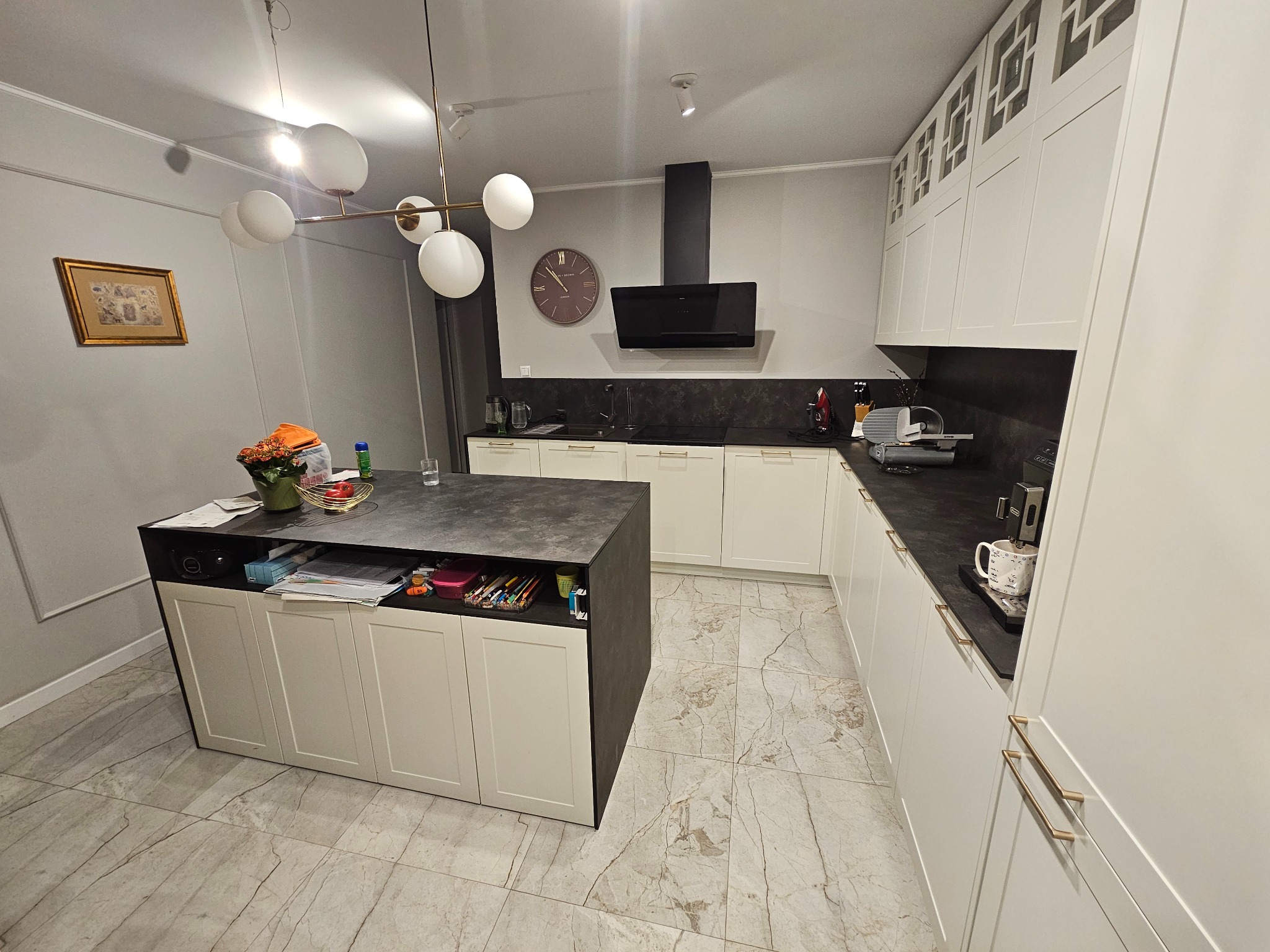 Nowoczesna kuchnia z wyspą: białe fronty szafek, czarny blat, marmurowe płytki podłogowe i designerska lampa sufitowa. Uwagę przykuwają detale, np. zegar na ścianie.