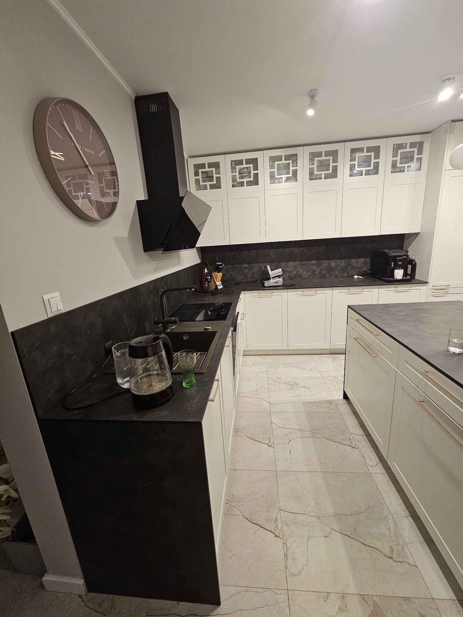 Nowoczesna kuchnia z białymi szafkami, czarnym blatem i okrągłym zegarem na ścianie. Widoczny zlew, czajnik elektryczny i marmurowe płytki podłogowe.