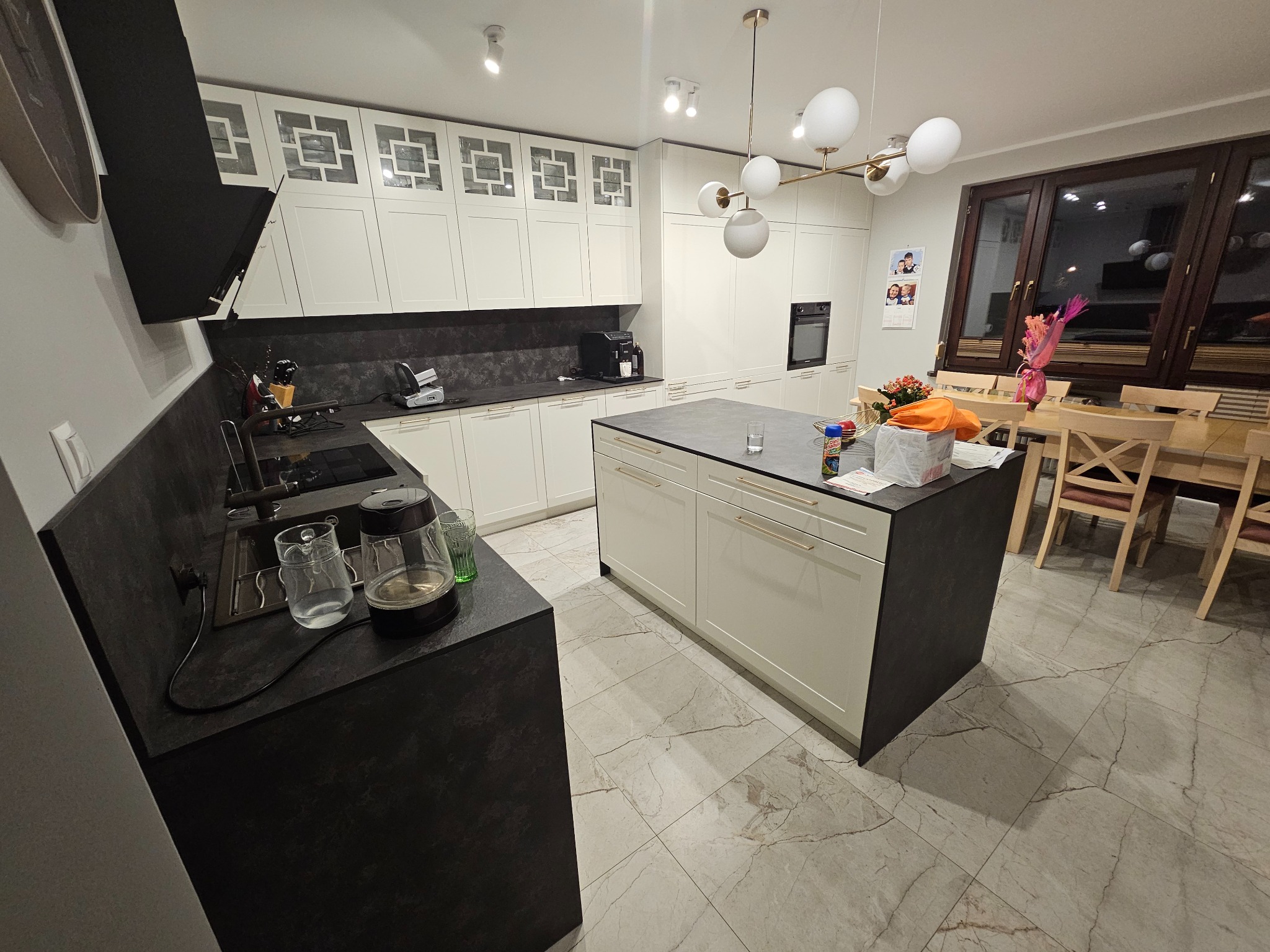 Nowoczesna, jasna kuchnia z wyspą i białymi szafkami z geometrycznym wzorem. Ciemny blat i marmurowa podłoga. Lampa sufitowa z kulistymi kloszami.