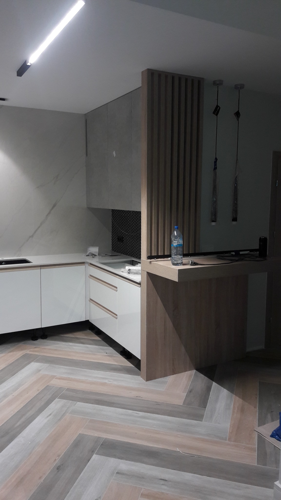 Nowoczesna kuchnia z białymi szafkami i drewnianą ścianką działową, oświetlona lampą sufitową. Na blacie butelka wody. Parkiet ułożony w jodełkę.