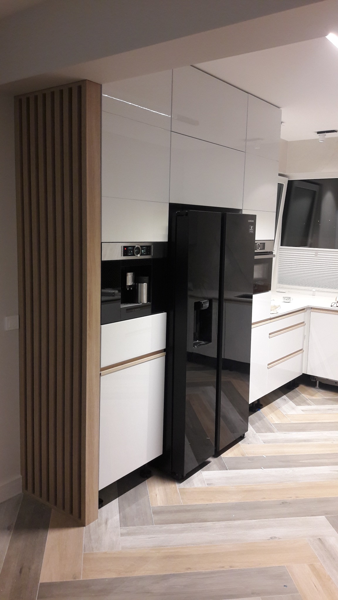 Nowoczesna kuchnia z białymi szafkami, zabudowaną lodówką i ekspressem do kawy, wykończona drewnianą lamelą. Parkiet w jodełkę na podłodze.