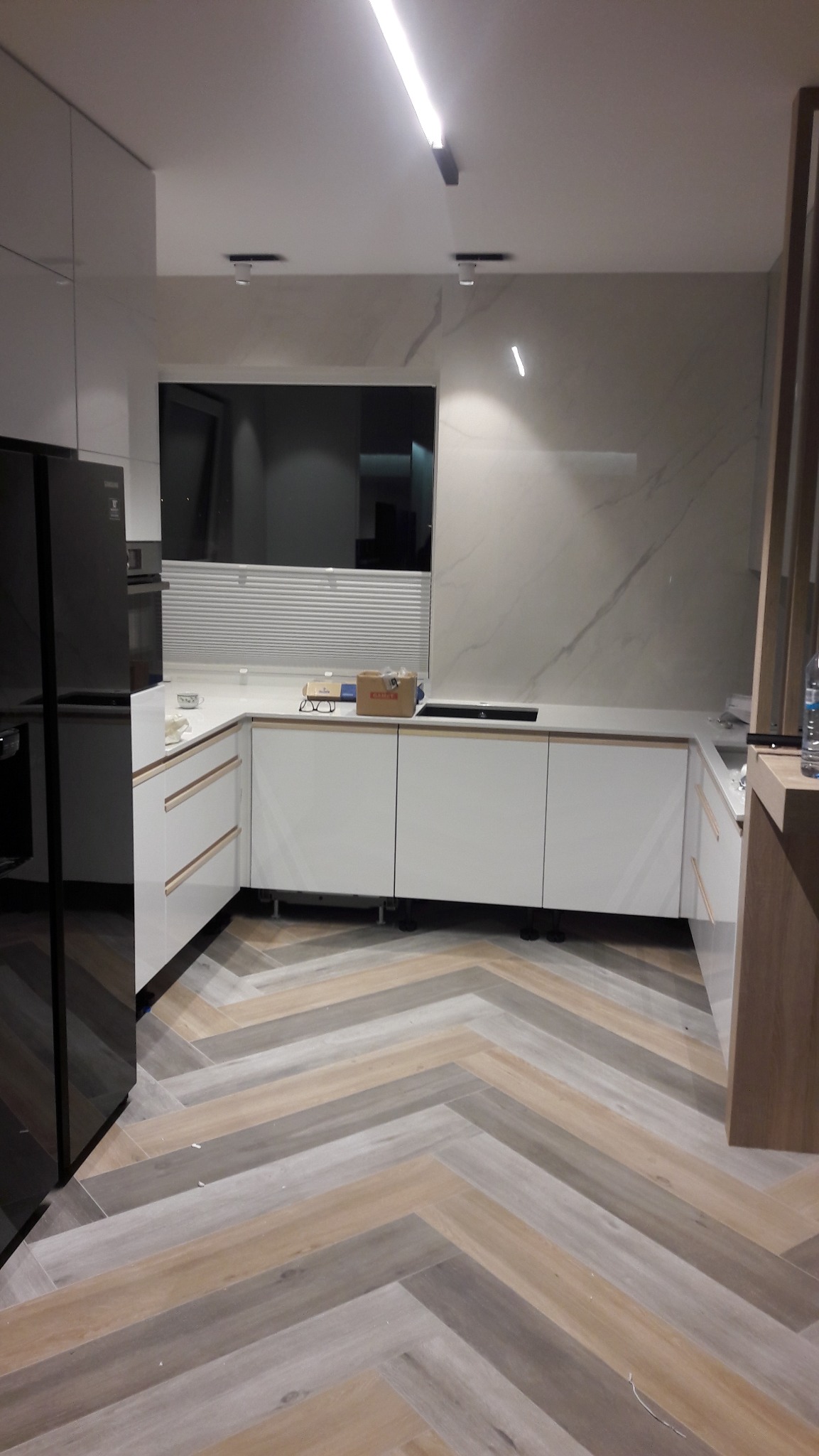 Nowoczesna kuchnia z białymi frontami i blatem w odcieniach szarości, podłoga ułożona w jodełkę, oświetlenie LED. Minimalistyczny design z czarną lodówką.