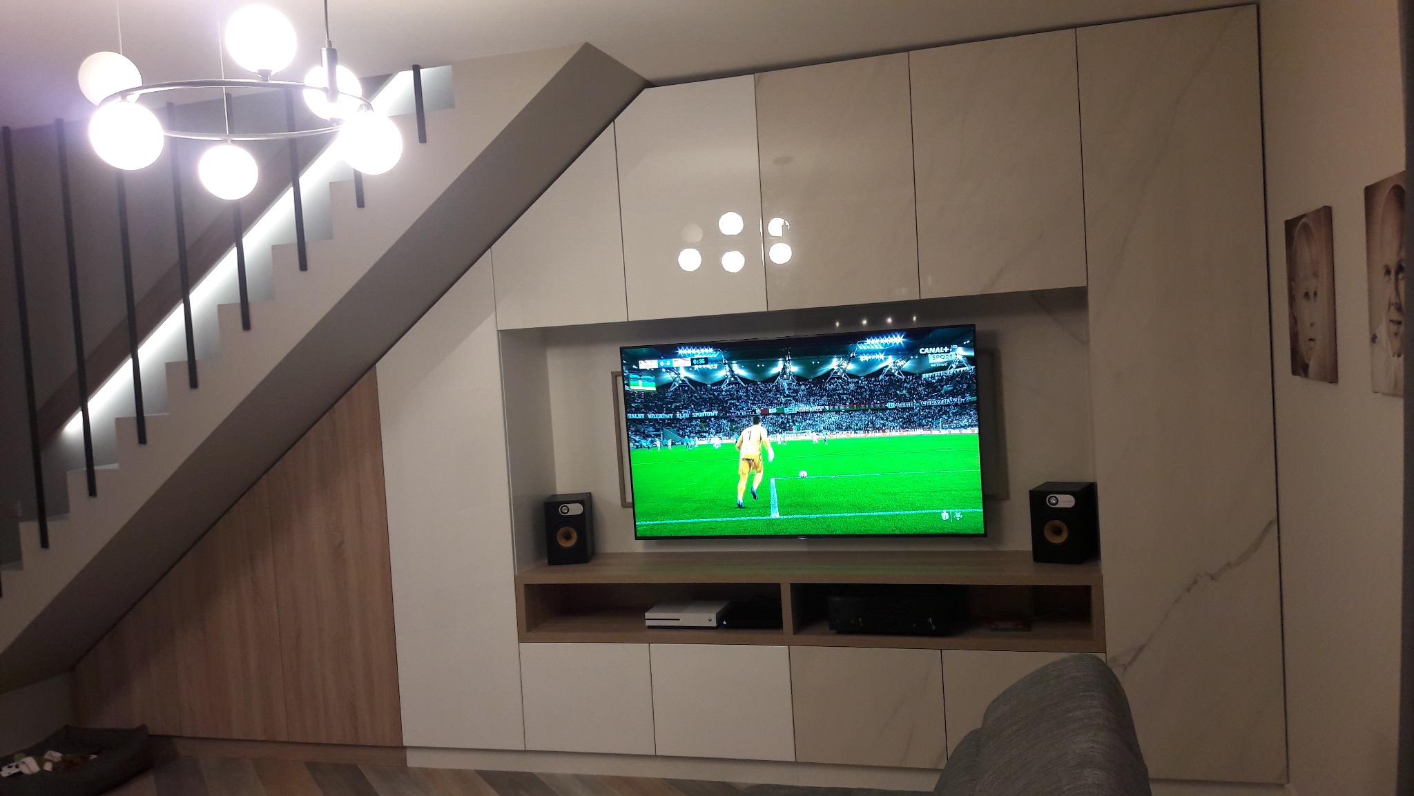Biała zabudowa meblowa pod schodami z szafkami, półkami i miejscem na TV. Nowoczesne oświetlenie schodów i sufitu.