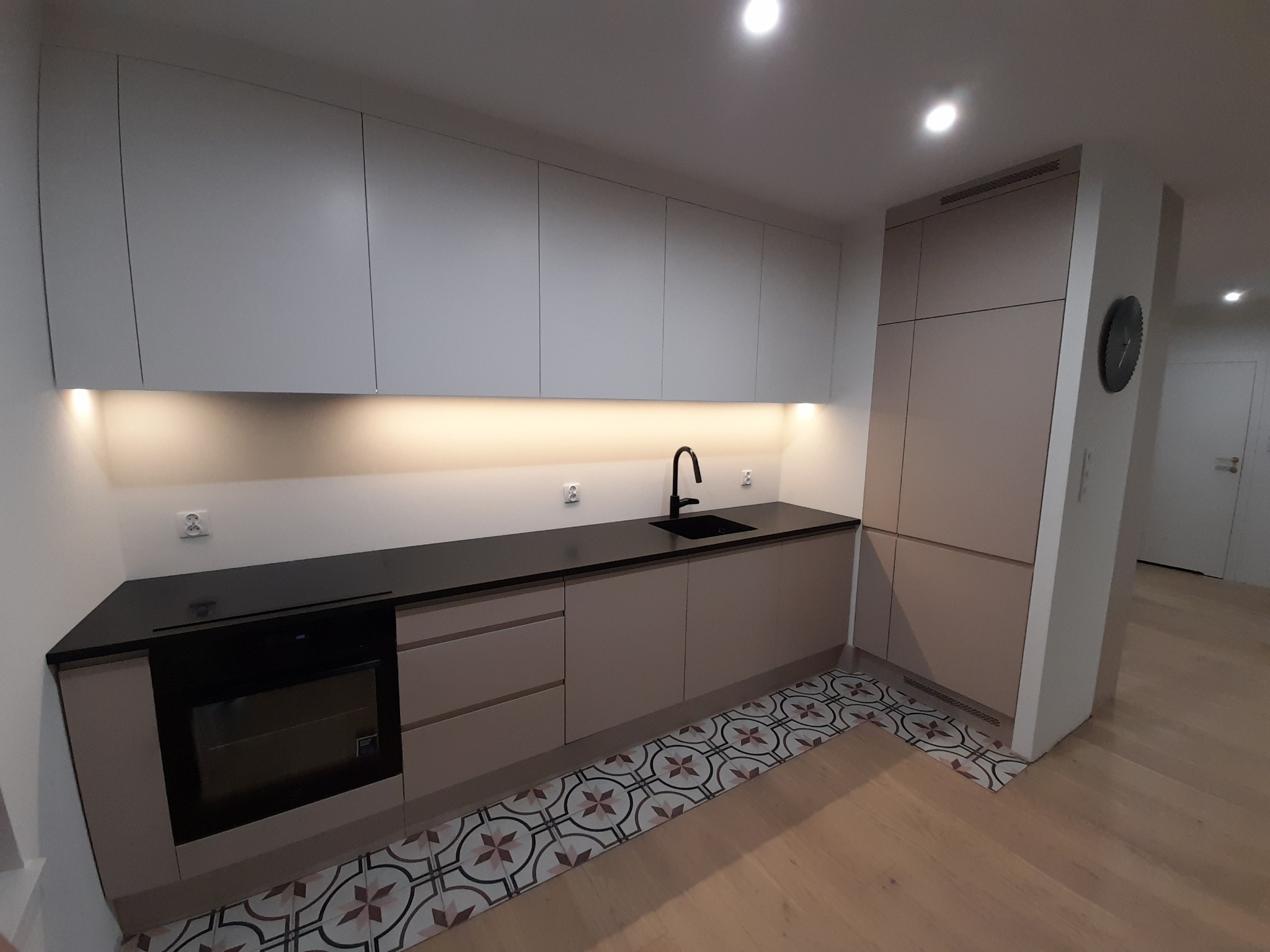 Jasna kuchnia z czarnym blatem, fronty w odcieniach beżu, wzorzysta podłoga. Nowoczesna aranżacja z oświetleniem podszafkowym i minimalistyczną armaturą.