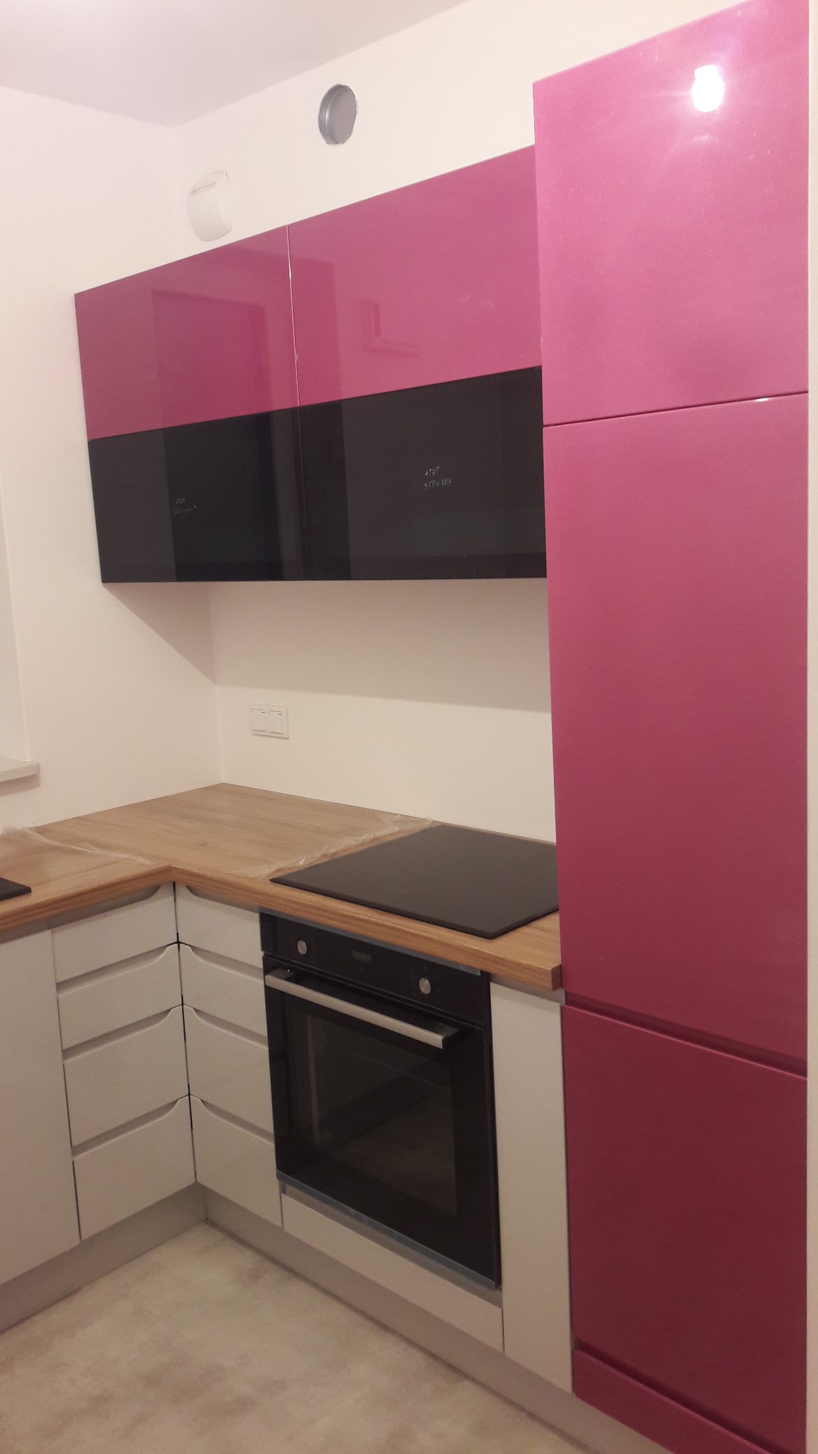 Nowoczesna kuchnia z różowymi szafkami górnymi i czarnym pasem, jasne blaty drewniane, piekarnik w zabudowie. Minimalistyczny design, oświetlenie punktowe na suficie.