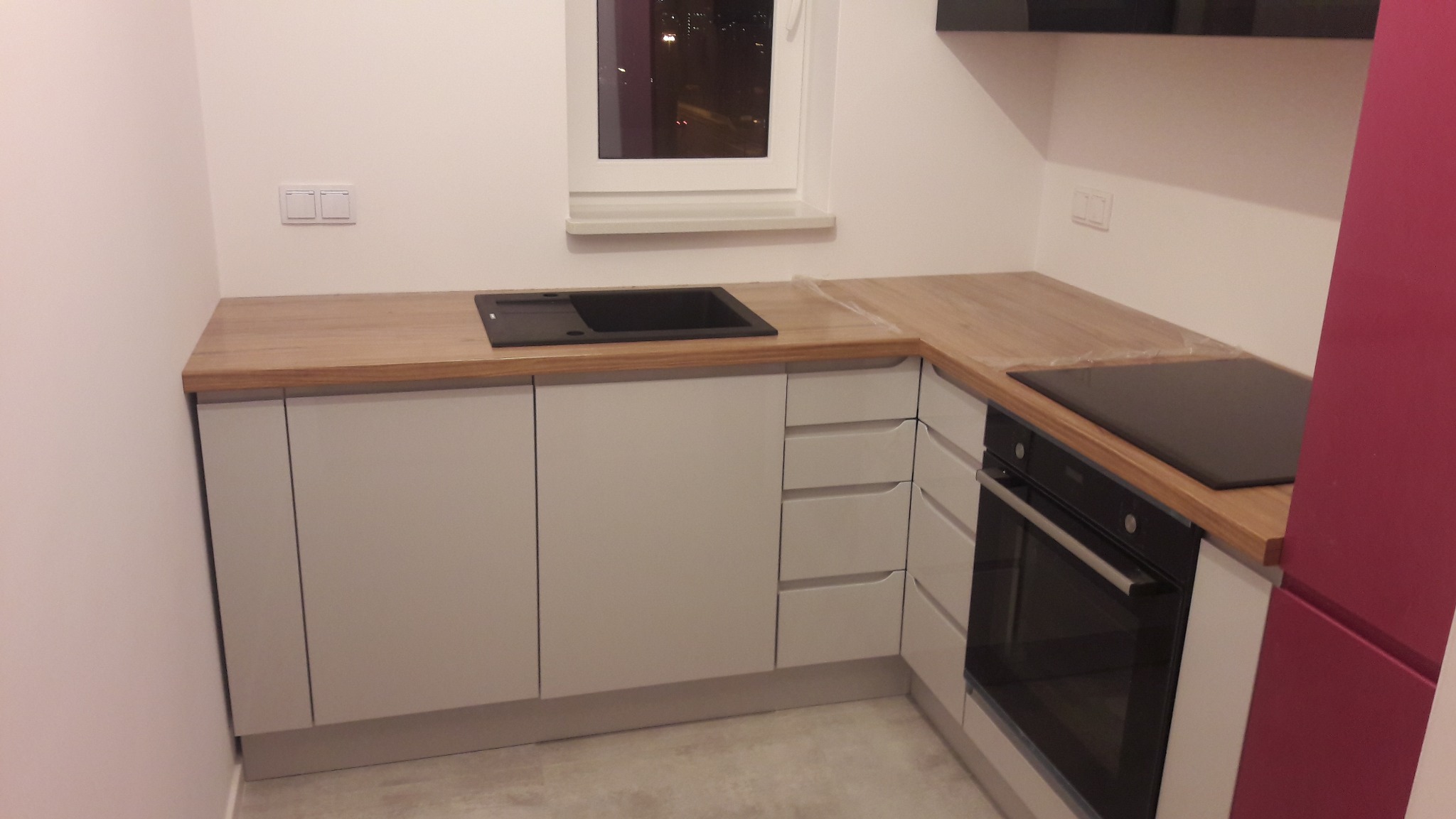 Jasna kuchnia z drewnianym blatem, czarnym zlewem i płytą grzewczą. Minimalistyczny design, okno z widokiem na nocne miasto. Nowoczesne wykończenie.