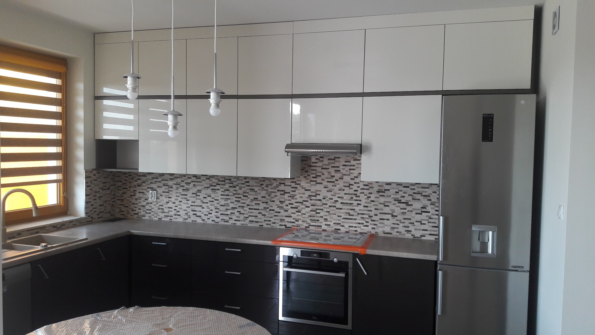 Nowoczesna kuchnia z białymi górnymi szafkami i ciemnymi dolnymi, mozaiką na ścianie i trzema wiszącymi lampami. Widoczny piekarnik i lodówka.